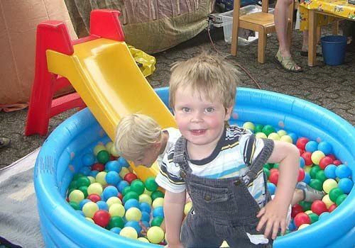 Kleiner Junge lächelt, während er aus einem Bällebad mit gelber Rutsche schaut.